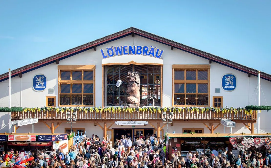 Exterior of Löwenbräu Festzelt at Oktoberfest with its iconic 4.5-meter roaring lion statue above the entrance, Bavarian flags, and crowds entering.