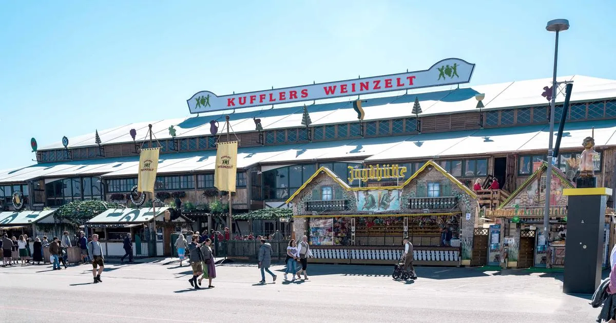 Kufflers Weinzelt tent exterior at Oktoberfest with rustic wooden facade, wine barrels, and guests toasting under string lights.