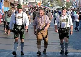 Three elderly men in traditional lederhosen, feathered hats, and embroidered suspenders walk joyfully, smiling and observing a lively festival scene.