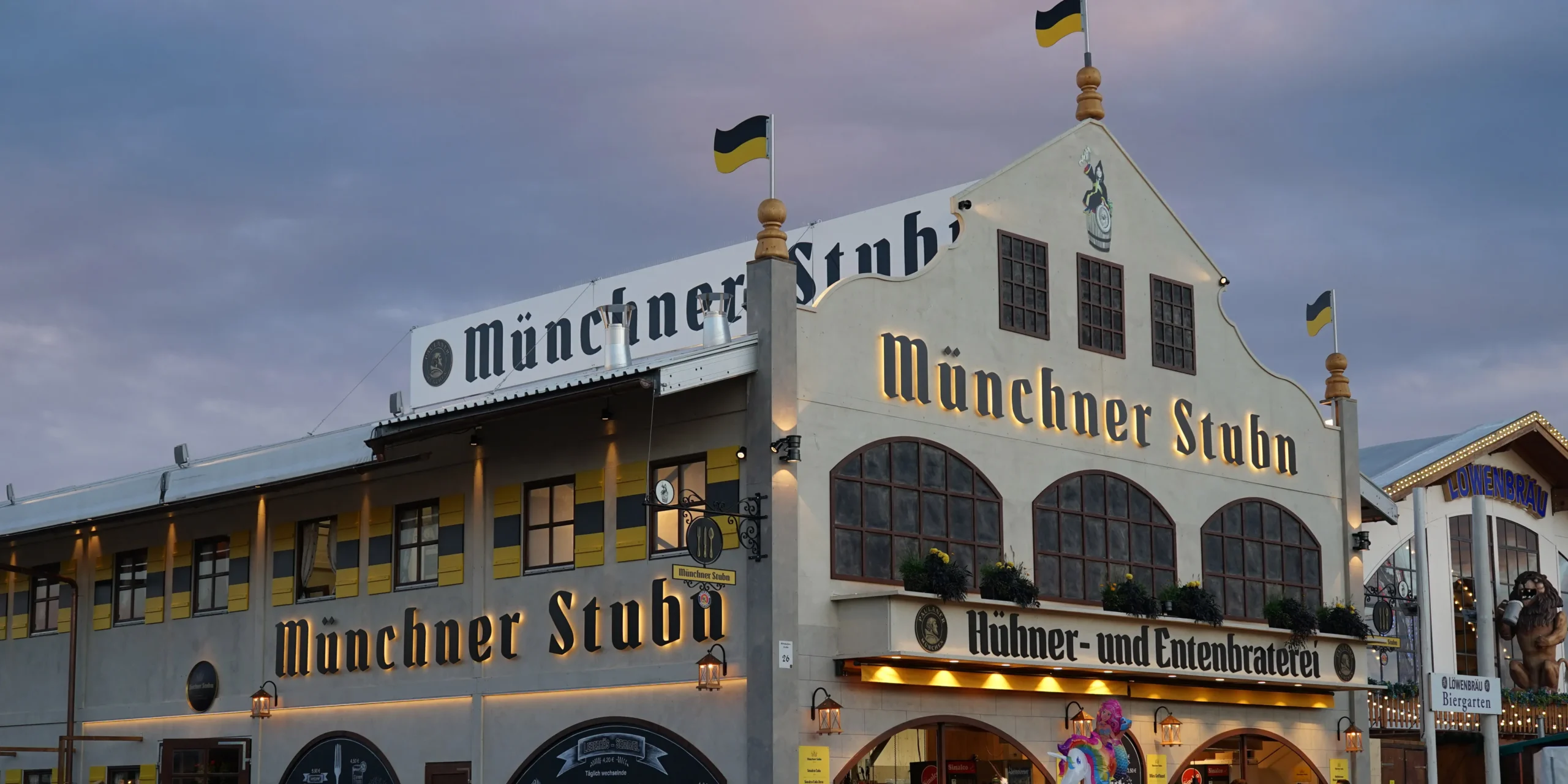 Rustic wooden sign for Münchner Stubn Oktoberfest tent with Bavarian typography, featuring repeated 'Münchner Stubn' text and 'Hilmer- und Entenbracher' specialty designation.