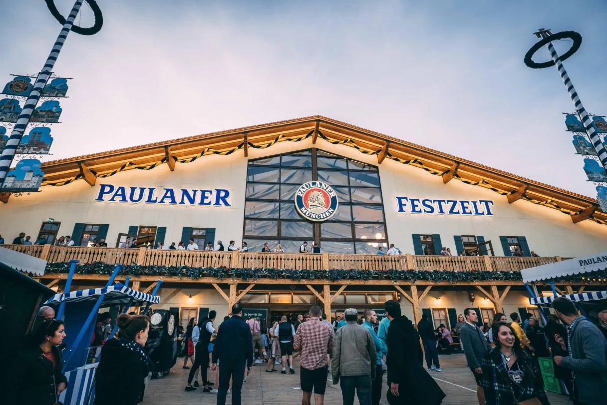 Paulaner Festzelt exterior with spinning beer tower, illuminated 'PAULANER FESTZEN' signage, and Oktoberfest crowds in traditional attire.