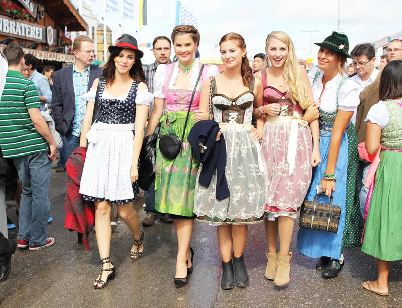 five young women in Bavarian dirndls with floral hairbands, crossbody bags, and ankle boots pose joyfully for a group photo.