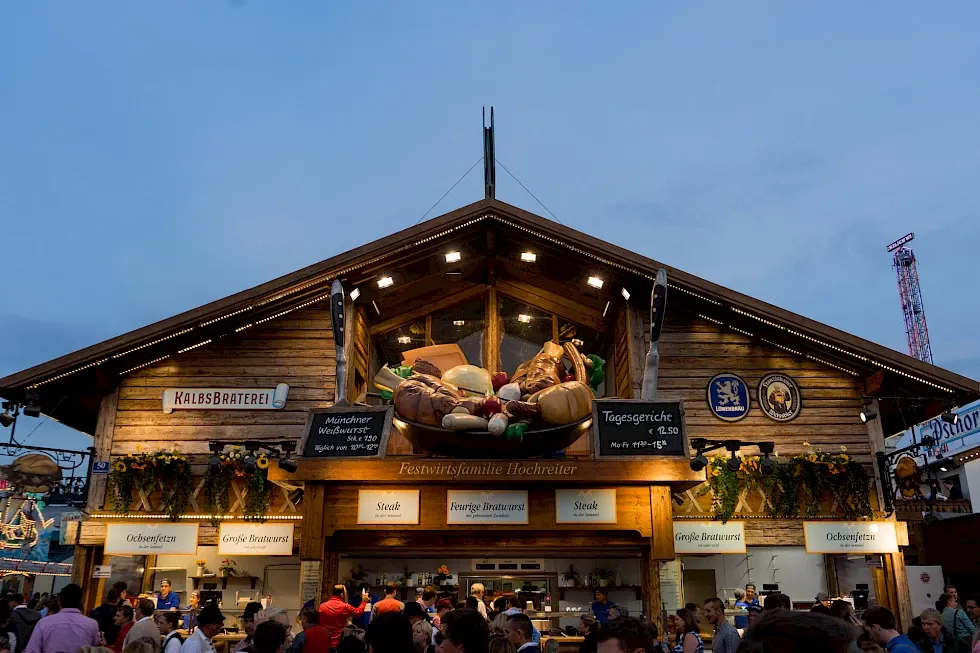Vintage-style sign for Kalbsbraterei Hochreiter veal roastery at Oktoberfest, featuring daily specials (Tagesgerichte) for €12.50 and terms like Mandrine and "Royal Arm 200-250" alongside grill imagery.