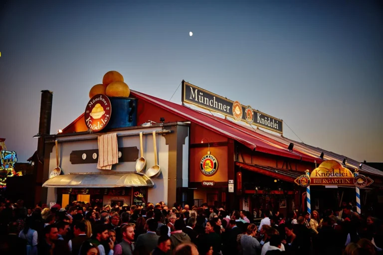Charming sign for Münchner Knödelei Oktoberfest tent featuring Bavarian dumplings (Knödel), with playful typography and Biergarten mention, set against rustic wood textures.